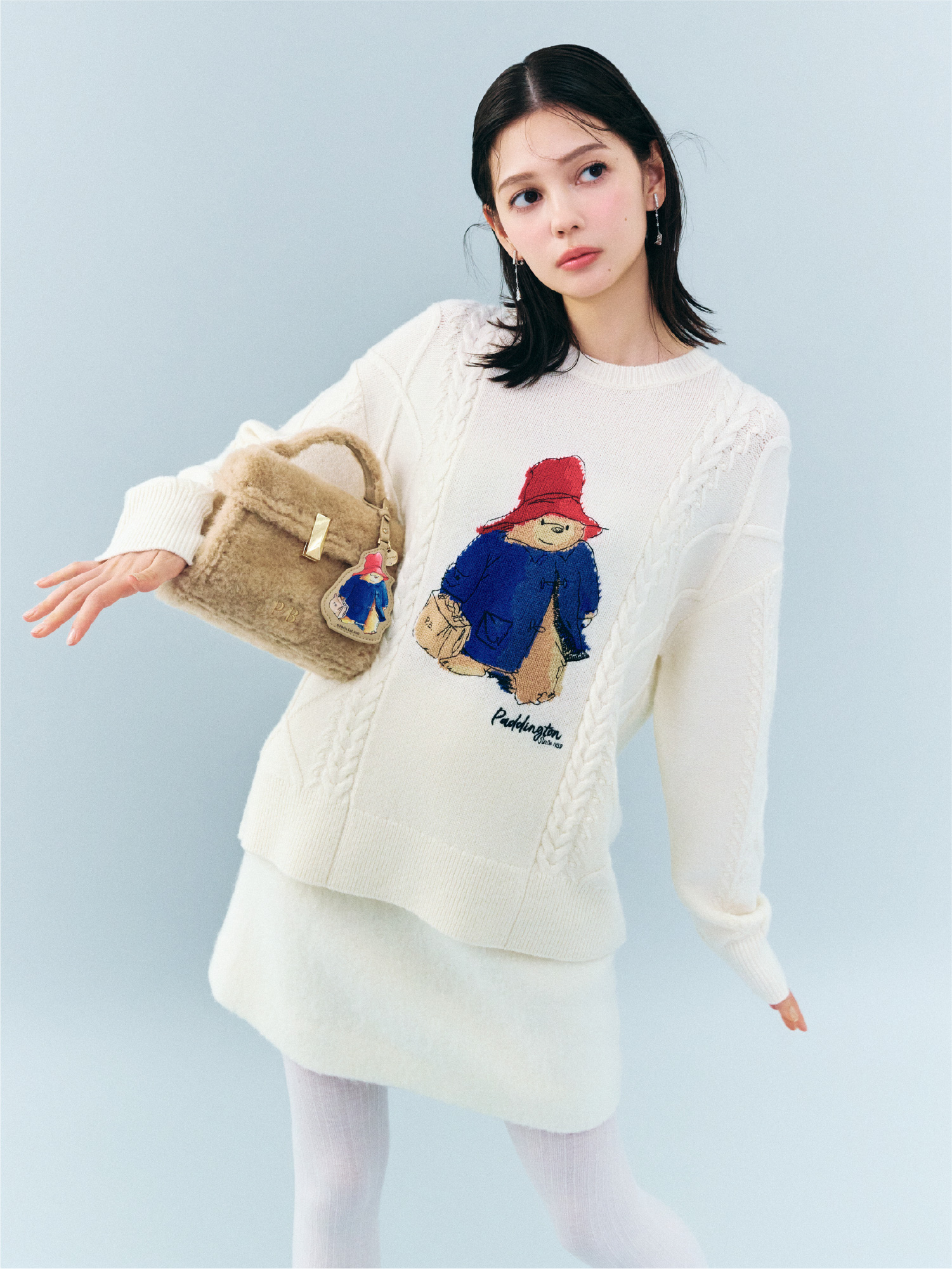 アイボリー色の【パディントン™×CELFORD】ケーブルニットプルオーバー、白色のバリエーションミニスカート、【パディントン™×CELFORD】ビジューハットイヤリング、【パディントン™×CELFORD】【リュタン】ビジュースタッズボアポシェットを着用したモデル画像