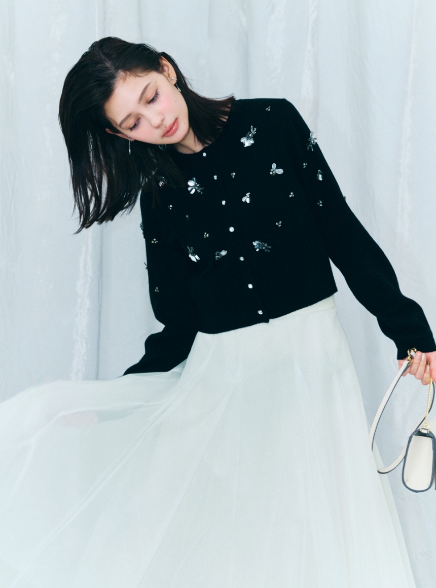 ブラック色のペタルビジューカーディガン、アイボリー色のシアーチュールスカートとビジューツイストバックルショルダーバッグを着用した女性のイメージ