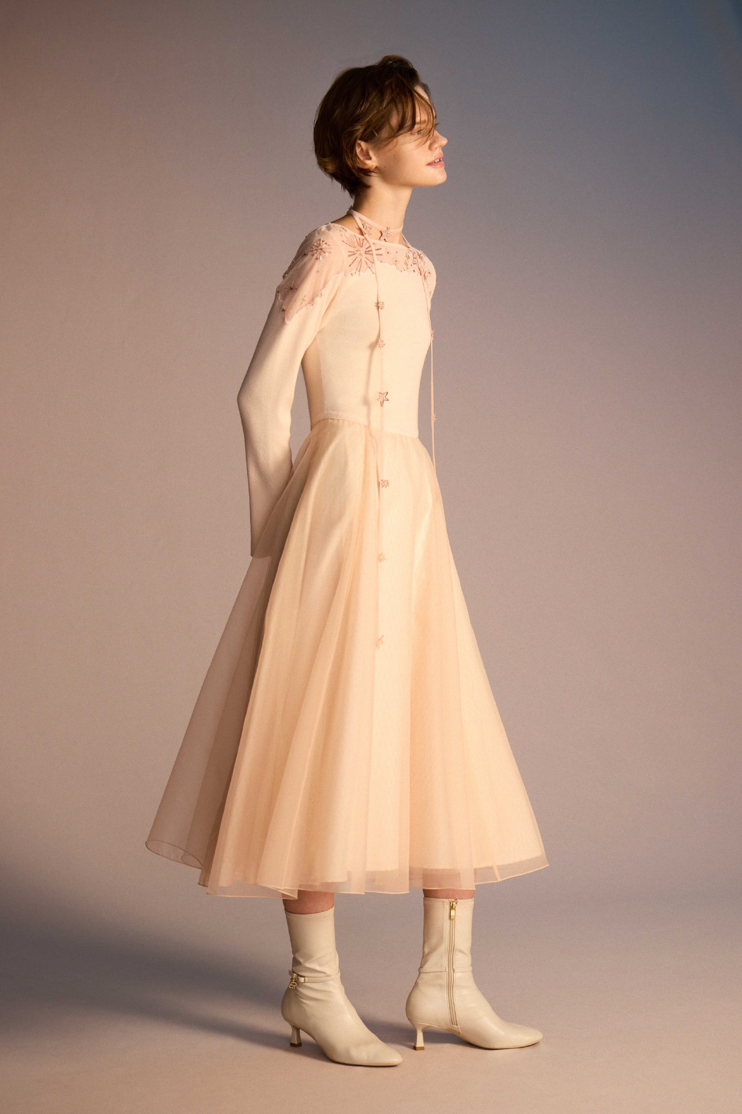 コーラル色のスターリーニットドレスとアイボリーのロゴチャーム付きストレッチショートブーツの着用例