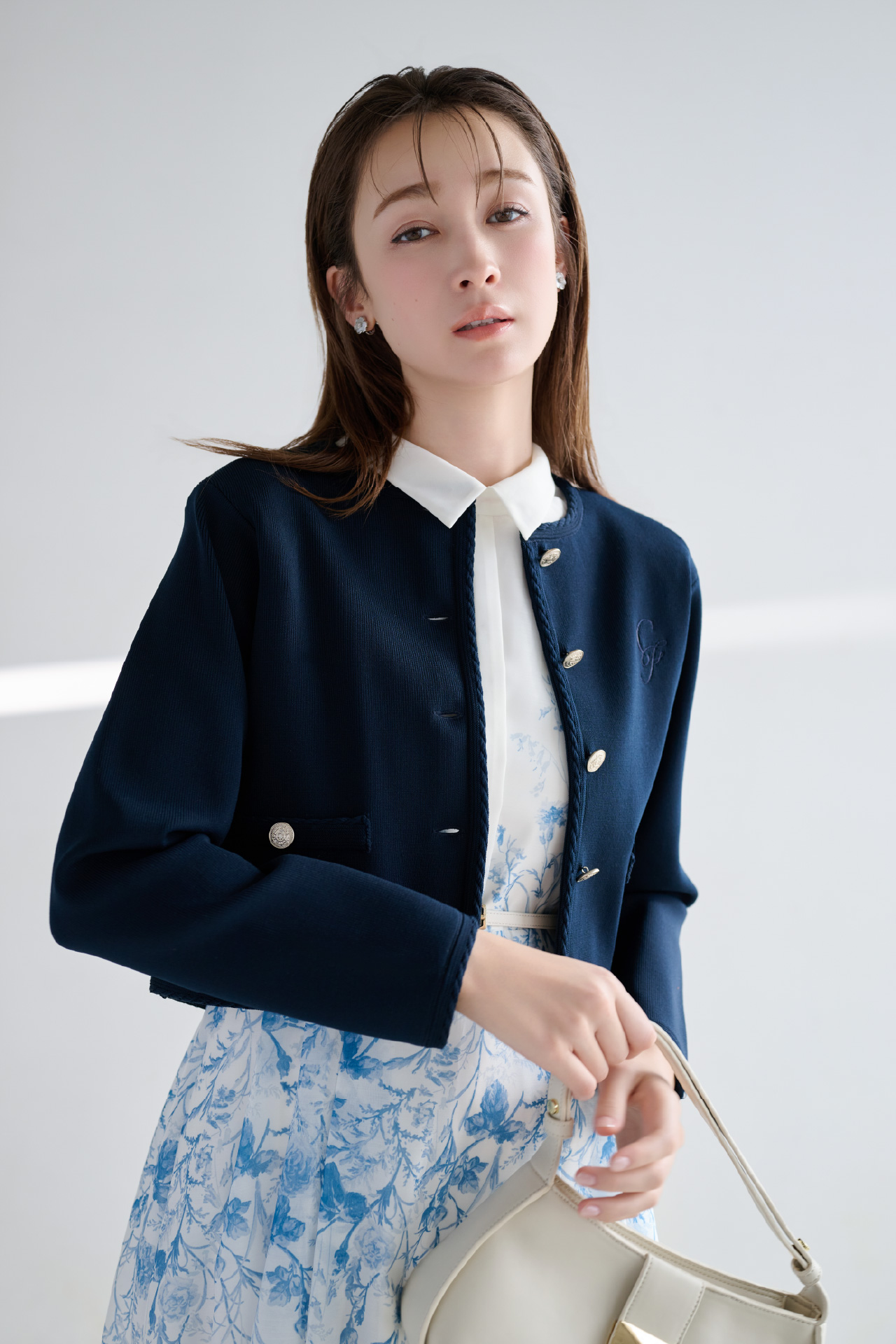 ネイビー色のトリム刺繍カーディガン、パネルフラワーワンピース、白蝶貝クローバーイヤリング、カリグラフィーチャーム付きナローベルト、2WAYワンハンドルショルダーバッグを着用した女性のイメージ